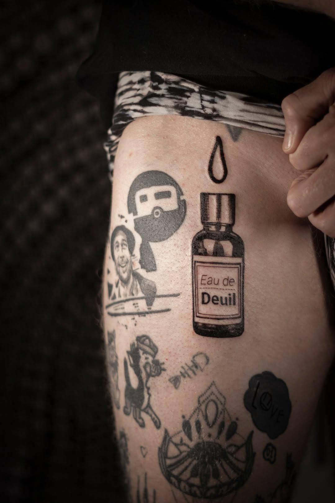 Multiple illustrative tattoos on thigh including Eau de Deuil bottle and figures